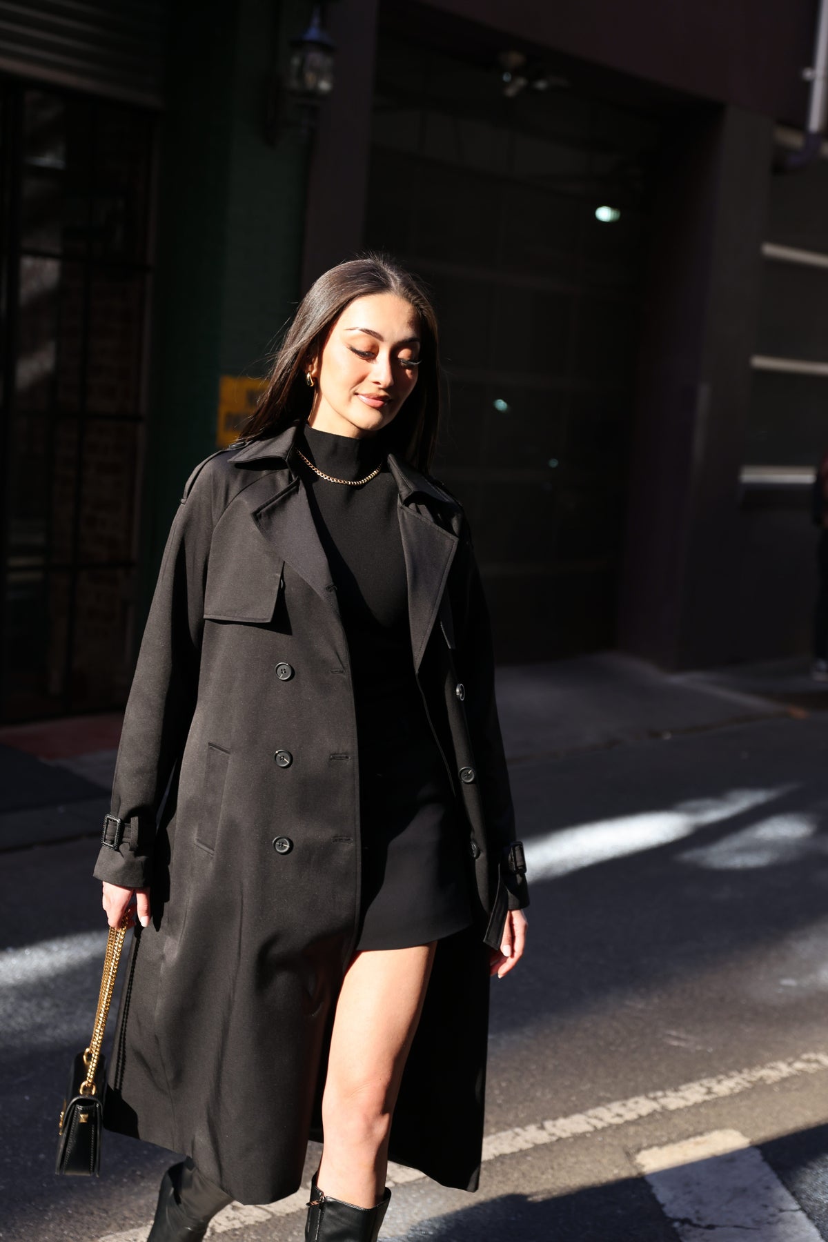 A woman wearing a long black trench coat over a black mini dress and black boots, walking on a city street in bright sunlight.