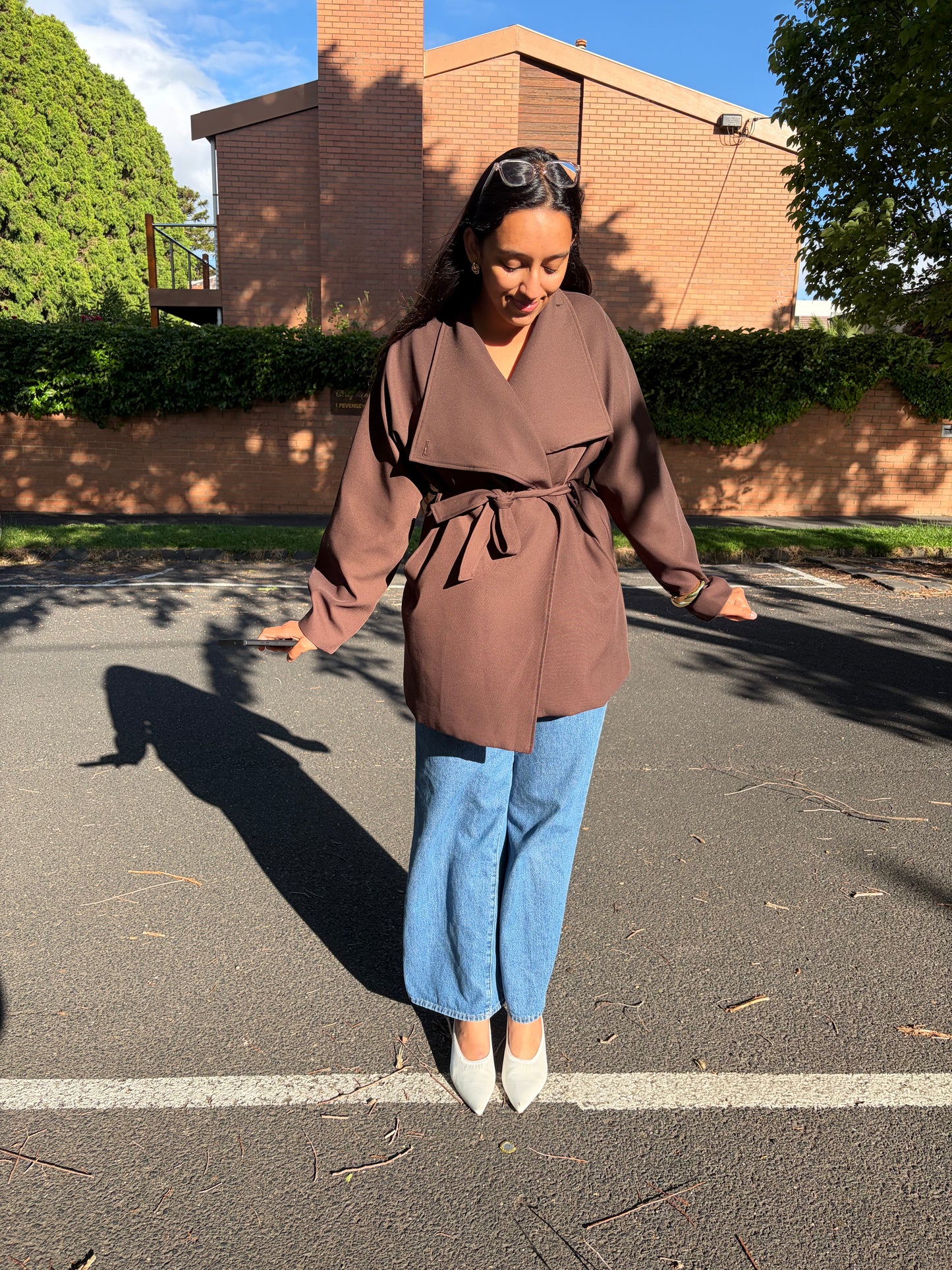 Woman wearing a brown coat and blue jeans standing on a street.