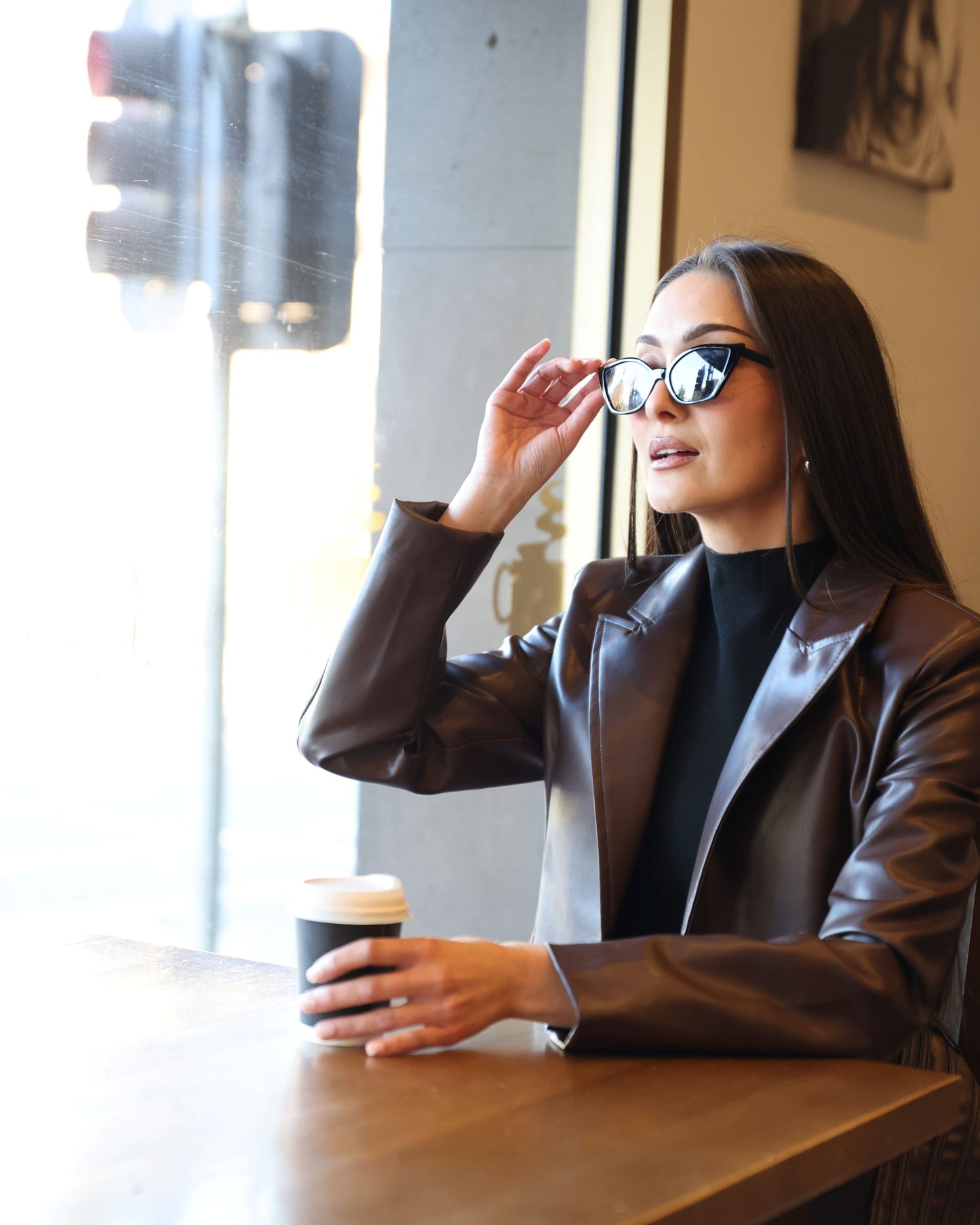 Woman sitting at a table with a coffee cup, wearing sunglasses and a leather jacket.