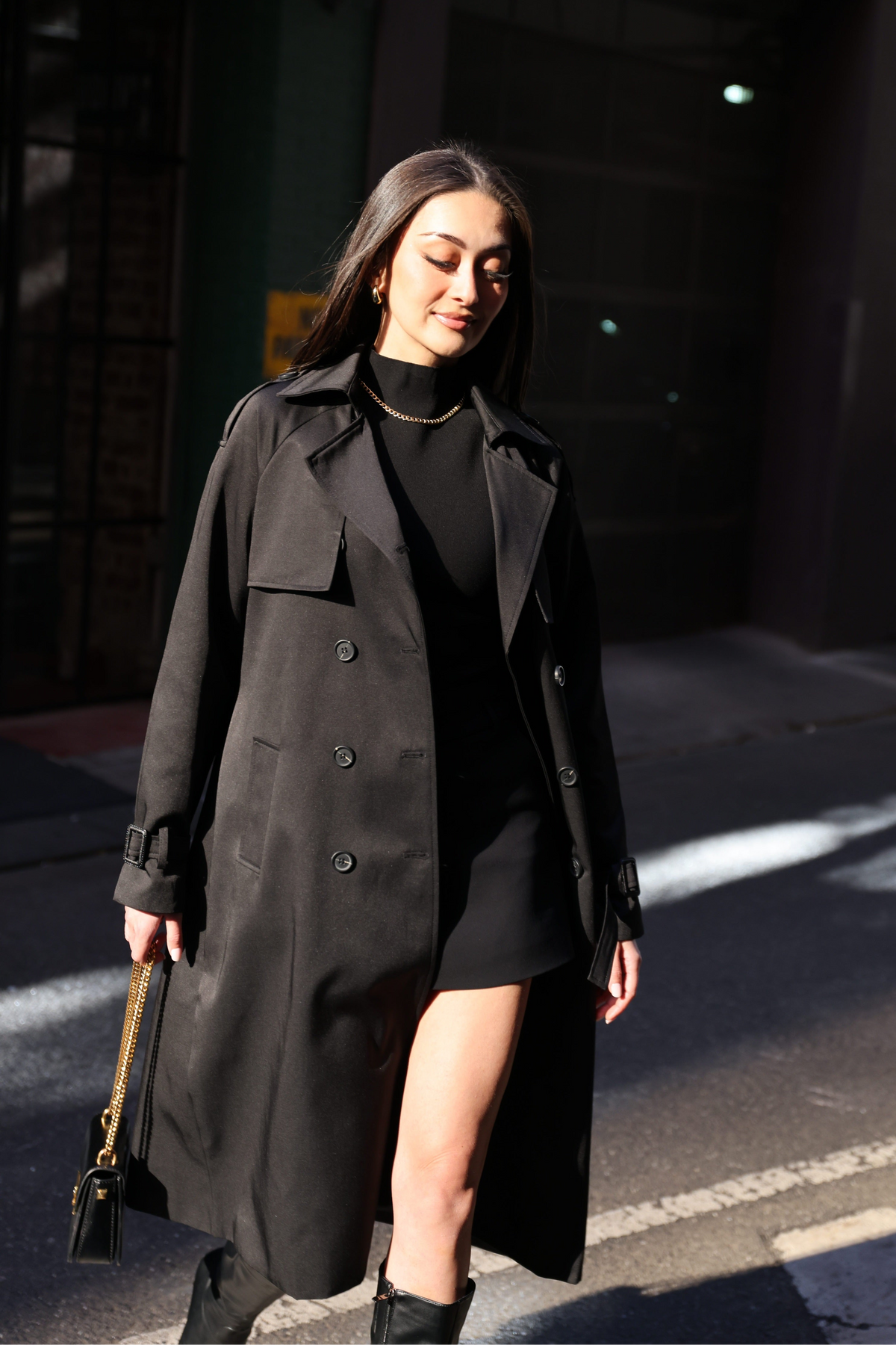 A woman wearing a long black trench coat over a black mini dress and black boots, walking on a city street in bright sunlight.